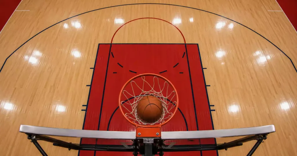 Indoor basketball court with the three-point arc and free-throw line clearly visible, viewed from above the baseline, ball passing through the hoop