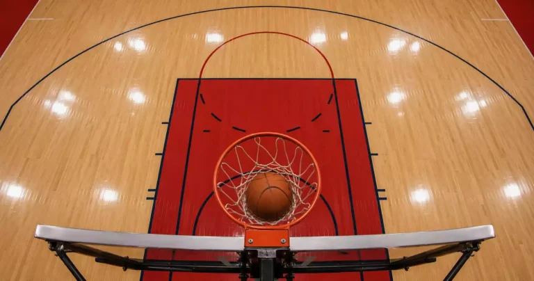 Indoor basketball court with the three-point arc and free-throw line clearly visible, viewed from above the baseline, ball passing through the hoop